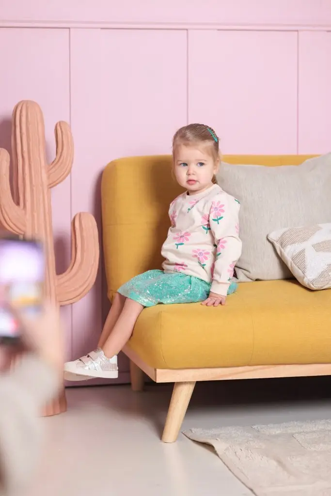 Een klein meisje zit op een gele bank in een kleurrijke daglichtstudio, met een roze muur en een cactusdecoratie op de achtergrond.