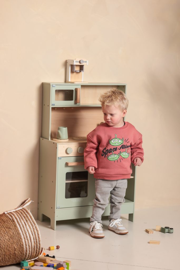 Een peuter in een roze trui met 'Space Alien' erop, speelt met een houten keukentje in een kleurrijke daglichtstudio.