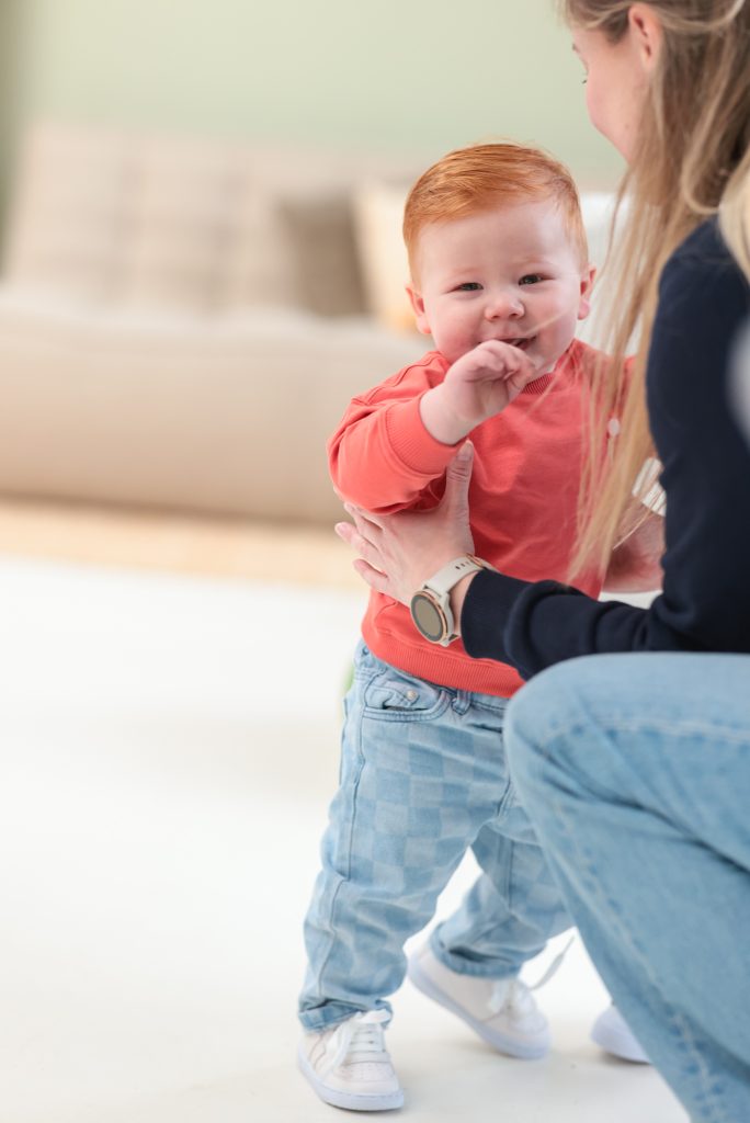 Een jonge peuter in een felgekleurde trui staat rechtop en lacht, terwijl een volwassene hem ondersteunt. De achtergrond is zacht en kleurrijk, passend bij een speelse fotoshootomgeving.