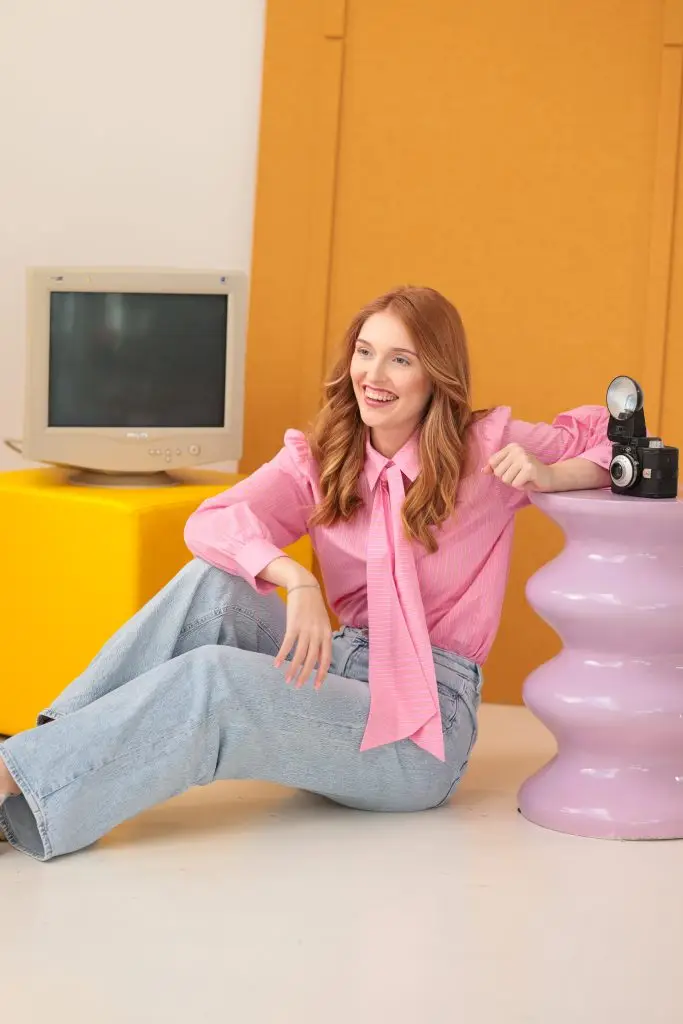 Vrouw met lange kastanjebruine haren, zittend op de vloer in een roze blouse met strik, en spijkerbroek. Achtergrond met een gele muur, een oud beeldscherm en een vintage camera op een paarse tafel.