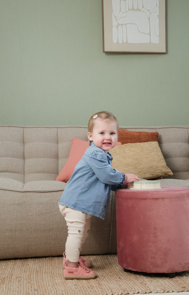 Een glimlachend peutermeisje staat naast een roze poef en voelt aan een speelgoedobject in een kleurrijke daglichtstudio.
