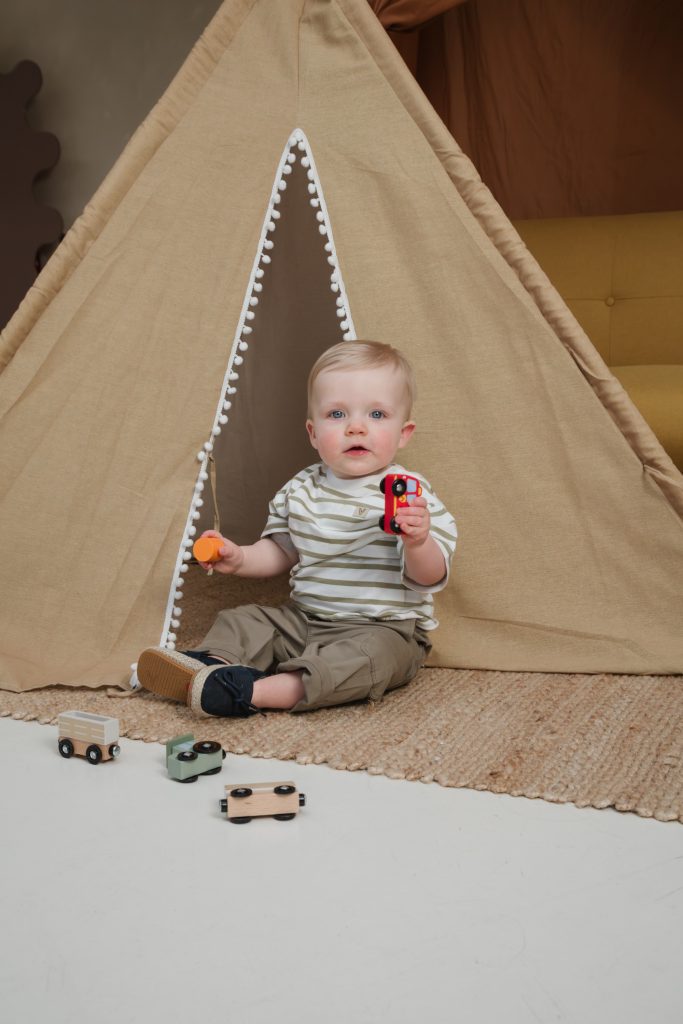 Een peuter zit voor een tipi-tent en houdt speelgoedauto's vast. Het kind draagt een gestreept shirt en zit op een vloerkleed met houten speelgoed om zich heen.