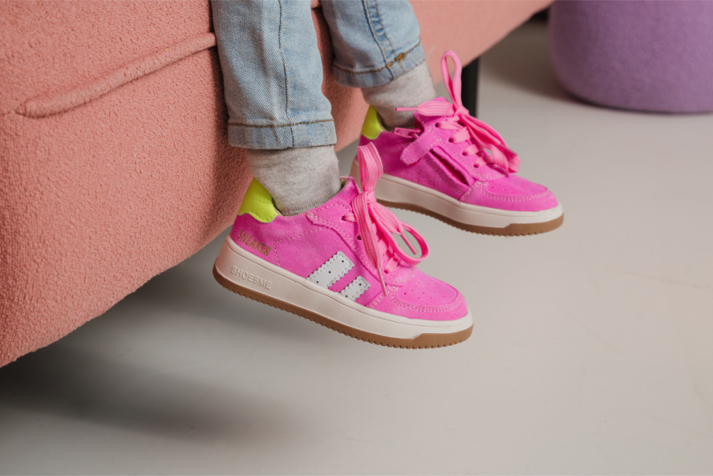 Een close-up van de schoenen van een kind, met opvallende felle roze sneakers met een groene hiel, terwijl het kind op een roze bank zit.