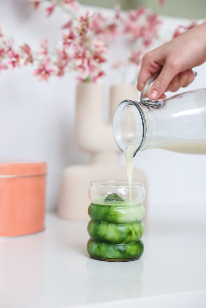 Een hand giet een groene drank uit een glazen karaf in een glas met groene en witte lagen, op een witte tafel met bloeiende takken op de achtergrond in onze daglicht studio in Brabant